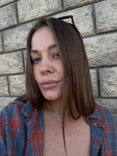 A young woman, with long brown hair and light skin, poses for a close-up photo. She is wearing a red and blue plaid flannel shirt. A delicate silver necklace with a circular pendant is visible around her neck. Her expression is neutral, with her eyes looking slightly to the side. She has dark eyebrows and light-colored eyes. A pair of black-rimmed glasses are perched on her head, partially obscured by her hair. The background consists of a textured stone wall, laid with rectangular blocks. The lighting suggests it is daytime. The image is oriented with the woman's head at the top. There is no visible text in the image.
