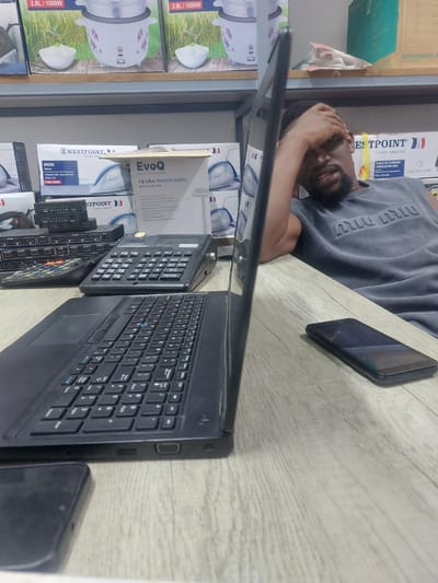 A man lies on a table covered in gray-white laminate, a phone next to his head and a laptop open in front of him. Numerous consumer goods such as electric kettles, irons, rice cookers, and microwave ovens are stocked on shelves adjacent to the table. The product brands visible include Westpoint, EvoQ, and Miu Miu. The location is most likely a retail store in Dar es-Salaam, Tanzania.