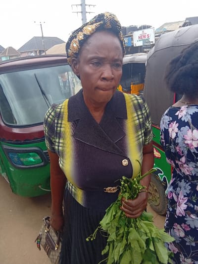 An adult female stands on an unpaved street in Ura Nta Umuarandu, Nigeria, with her body angled slightly to the right, looking forward. She wears a black and gold patterned headwrap, a short-sleeved top with a black central panel and vertical yellow and white striped sides, a black pleated skirt, and a gold-colored buckle/clasp at her waist. She holds a brown, patterned handbag with a striped strap in her left hand and a bunch of fresh green leafy vegetables in her right hand. Her lips are pursed.
To her immediate right, the upper torso and back of a second individual are visible, wearing a dark blue dress with a light pink and white floral pattern. Behind the primary subject and to the left, a green and red three-wheeled auto-rickshaw is partially visible. Further in the background, another dark-colored auto-rickshaw is present, along with buildings, including one with a white sign displaying "HEM'S D CHEMIST & SUPERMARKET". Utility poles are visible against an overcast sky. The ground appears to be dirt or gravel.