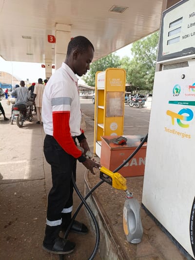 The image displays a TotalEnergies fuel station in Gawuna, Nigeria. A male fuel attendant stands with his body angled to the right, dispensing liquid from a yellow fuel nozzle into a grey plastic container positioned on the ground. He wears a white collared shirt with red and grey panels on the sleeves, black trousers with reflective strips, and black slip-on sandals, directing his gaze downwards at the container. To his right stands a white fuel dispenser, featuring a digital display indicating "NGN / Litre," along with "TotalEnergies" and CAF branding. A red payment terminal rests on a brown metal box next to the dispenser. Behind the attendant, a yellow display stand labeled "SOLAR ENERGY SOLUTIONS" is visible. The scene is under a white canopy supported by pillars, one marked with the number "3." In the background, several individuals and motorcycles are present, with trees and a bright sky in the distance. The ground is paved concrete.