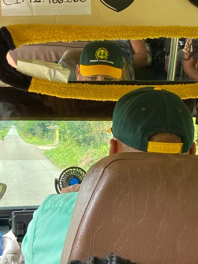 The image appears to be taken from inside a vehicle, looking forward through a windshield or window. In the foreground, the back of a person's head is visible, wearing a dark green baseball cap with a yellow brim and an emblem. The person is seated in a brown leather seat. To the left, a small black fan with a blue center is mounted.
The reflection in the mirror above shows another person wearing a similar dark green baseball cap with a yellow brim and an emblem. This person's face is partially visible, looking downwards. There are some papers or documents on a surface reflected in the mirror. A fluffy yellow and black trim borders the top of the mirror.
The environment outside the vehicle is a paved road stretching into the distance, lined with lush green vegetation. A motorcycle is visible on the road in the left distance. In the middle distance, there is a yellow pole structure with a blue sign on top, next to the road. The scene suggests a rural or semi-rural setting.
The text at the top of the image, partially visible, reads "C.1. 12.100.260". Based on the context of San José de Barlovento, Venezuela, this could be a registration number or identifier. The overall impression is a candid shot of individuals traveling in a vehicle, possibly a bus or van, in a tropical or subtropical environment.