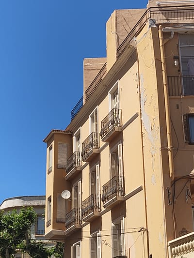 The image shows a section of a building in Dénia, Spain. The building is a multi-story structure with a warm, light yellow facade. The paint on the facade is peeling in several areas, showing signs of weathering and age. Each story features windows with white shutters and small balconies with ornate black iron railings. A satellite dish is attached to the side of the building near one of the windows. A dark metal railing is present along the top edge of the building. A drainpipe runs vertically along the right side of the building. In the background, the clear blue sky contrasts sharply with the building's color, and parts of other buildings and trees are visible.