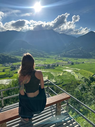 A female, viewed from behind, is seated on a wooden bench part of a viewing platform overlooking a vast agricultural valley in Tú Lệ, Vietnam. She has shoulder-length brown hair and wears a dark green crop top with a dark jacket draped around her waist. A colorful, intricate tattoo covers her left shoulder and upper arm. The platform features metal railings and a corrugated roof structure beneath the bench.

The expansive valley below is dominated by vibrant green terraced rice paddies, some fields containing water that reflects the sky. Small villages with houses are interspersed throughout the fields, and a winding body of water, likely a river or stream, is visible. In the background, majestic, hazy blue-grey mountains frame the valley under a bright blue sky with scattered white clouds. A prominent sun flare emanates from the upper left, casting strong light across the scene, and a faint rainbow-like optical phenomenon is visible near the sun.