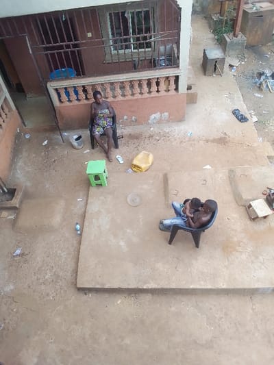 The image depicts an outdoor residential area in Ukwu-Abwa, Nigeria. The scene is dominated by a multi-tiered dirt and concrete ground surface.
**Environment:** To the left, a two-story building with a reddish-brown facade and a visible balcony on the upper floor features a brown metal railing and orange concrete balusters. Windows with security bars are present on the upper level. The ground consists of irregular patches of concrete and packed earth, with numerous small pieces of refuse, including plastic bottles and wrappers, scattered throughout. In the mid-ground, a larger concrete platform is visible. The background to the right shows a cluttered area with disturbed earth, ashes, and small, cubic structures, suggesting a refuse burning or outdoor activity zone.
**People:**
1. **Female:** On the left, an adult female with dark skin is seated barefoot in a black plastic chair, wearing a patterned, multi-colored garment. Her white flip-flops are on the ground nearby. A clear plastic bucket is positioned near her feet.
2. **Male & Child:** On the right-center, a shirtless adult male or older adolescent with dark skin is seated in a black plastic chair, wearing light-colored jeans. He is holding a young child, possibly an infant or toddler, who is partially clad in a light-colored garment. Both are oriented towards each other, appearing to interact.
**Objects:** A small, light-green plastic stool is situated between the female and the male figures. A yellow plastic jerrycan lies on the ground to the right of the female. A light-colored, circular plate or disc rests on the concrete platform in the central area. A discarded dark garment is visible on the far right ground.