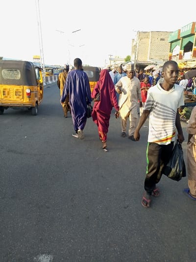 The image shows a street scene in Maiduguri, Nigeria, taken at the coordinates 11.831, 13.154. A group of people are walking on a paved street. The people are dressed in traditional clothing, including a woman wearing a long red dress and headscarf, a man in a blue robe, and a man in a white robe. There are two yellow auto rickshaws in the background. The street appears to be in an urban area. The background shows buildings with signs and advertisements. The image shows a bustling scene with people moving about on a busy street. The image was likely taken during the day as the sun is bright and the light is strong.