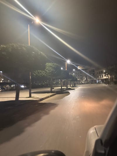 A nighttime street scene is captured from within a vehicle, showing a road illuminated by streetlights. The lights cast a strong glare, creating starburst patterns. Buildings line the left side of the road, interspersed with trees casting dark shadows. The pavement is visible, along with portions of the vehicle, including a tire. The overall scene is dark and suggestive of motion.