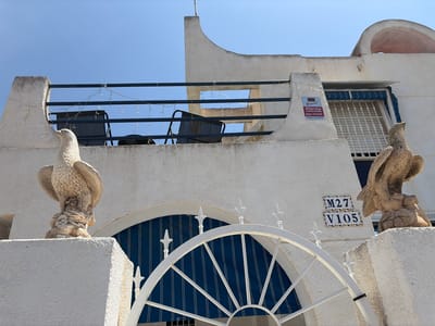 This is an eye-level, medium shot of a white stucco building in Torrevieja, Spain. The building has a balcony with dark metal railings decorated with small, white string lights. Two dark chairs are visible on the balcony. Two stone eagle statues are positioned on white stucco walls in the foreground. One statue is on the left, facing left, and the other is on the right, facing right. Between the statues, a white metal archway is partially visible, leading to an arched entryway with blue and white striped awnings and a barred window. Two address tiles are mounted on the building wall: "M27" above "V105". A red and white security sticker for "DIRECT ALARMA" is also on the wall to the left of the window. The sky is clear and blue.