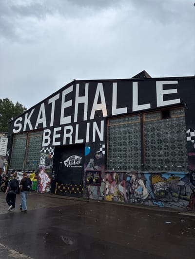 The image shows the "Skatehalle Berlin," a well-known indoor skatepark located at Revaler Straße 9, Berlin, Germany. The building features large white lettering across its black facade that reads "SKATEHALLE BERLIN." The entrance is a black gate with a "Vans Off The Wall Skateboarding" logo. The exterior walls are heavily decorated with graffiti and murals. In the foreground, two individuals are walking away from the camera, one wearing a black backpack and the other a brown backpack. The ground is wet and appears to be a metal grate or textured surface, reflecting the overcast sky. The overall environment suggests an urban setting with a focus on skateboarding culture.