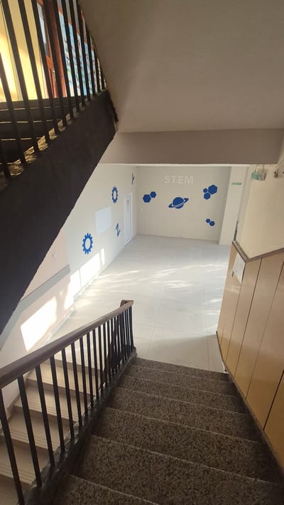 The image shows an interior view of a building's staircase and landing area, possibly in a school or educational facility. The scene is captured from a descending perspective on a concrete staircase with a dark metal railing. The landing area below features a light-colored tiled floor and pale beige walls. The walls are decorated with blue decals, including hexagonal shapes, gears, a planet with rings, and the word "STEM" in large, block letters. There is a white board on the wall and a door. An "EXIT" sign is visible above the door. Sunlight streams in from a window on the left, casting shadows on the wall and floor. The architecture suggests a modern but possibly older building, with a prominent dark wooden banister on the stairs. The overall impression is one of a functional educational space. No people are visible in the image.