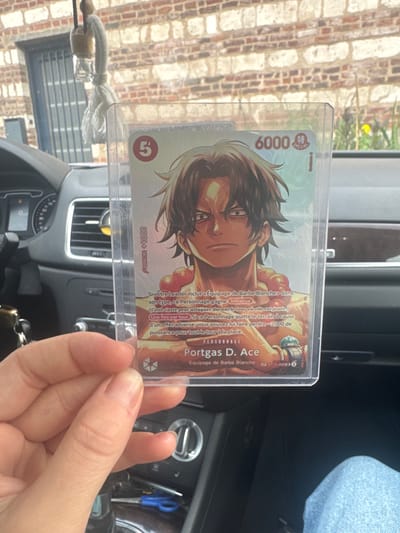 In Villeneuve-d'Ascq, France, a person is holding a transparent plastic-sleeved trading card featuring the character Portgas D. Ace from One Piece. The card has an illustration of Ace with a determined expression, wearing a red beaded necklace and a green band on his left arm. Text on the card is visible, including "6000", "CONTRE +1000", and "Portgas D. Ace". The scene appears to be inside a car, with the dashboard and a brick building visible in the background through the car's windshield. The person's hand, with fingernails visible, is holding the bottom edge of the card.