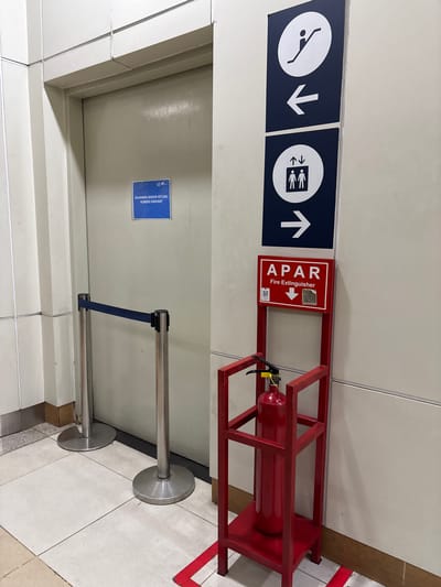 The image shows a hallway in what appears to be a public building, possibly an airport or train station, given the signage and crowd control barriers. The setting is indoors, with tiled flooring and light-colored walls.
The primary subjects are directional signs and a fire extinguisher. On the left, there are two dark blue signs with white pictograms and arrows. The first sign shows a person in a diving pose with a downward arrow, indicating a downward direction. The second sign depicts multiple figures with upward and downward arrows, suggesting movement of people in both directions. To the right of these signs, a red metal stand holds a red fire extinguisher labeled "APAR Fire Extinguisher" in white text, with a downward arrow and a QR code.
In the foreground, two silver stanchions with blue retractable belts are set up, suggesting a queue or restricted access area. A door is visible on the left behind the stanchions, with a blue sign attached to it. This sign contains text that appears to be in Indonesian, with a warning that access is prohibited except in emergencies.
The overall scene is one of an organized public space with clear indications for movement and safety equipment. The lighting suggests it is daytime, but no external weather conditions are visible. The location cues, such as the "APAR" label (common in Indonesia for fire extinguishers) and the Indonesian text on the door sign, point to this being in Central Jakarta, Indonesia. There are no people actively engaged in any specific activity, but the presence of directional signs and barriers implies ongoing foot traffic or controlled entry.