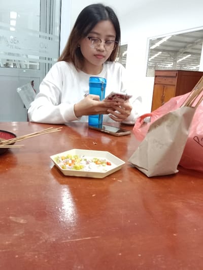 A young woman with glasses is seated at a table, occupied with a mobile phone. On the table, there is a small white hexagonal dish containing chopped vegetables, possibly a condiment. A blue water bottle and a second mobile phone are beside her. A brown paper bag and a red plastic bag containing satay skewers also sit on the table. In the background, there's a glimpse of an interior space and a wooden cabinet.
