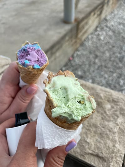 A first-person, close-up view shows a hand holding two ice cream cones. The cone on the right contains green ice cream and is wrapped in a white paper napkin. The cone on the left holds purple and blue ice cream. The hand has light skin and a fingernail painted light purple. A black and white object is visible between the hand and napkin. The background includes a light gray, textured surface, likely a stone wall or walkway. The location is likely in Olginka, Russia.