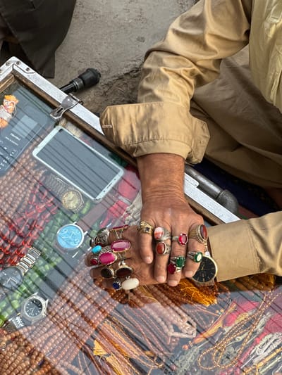 In Quetta City Tehsil, Pakistan, a person's hands are adorned with multiple rings, each featuring large, colorful gemstones, including red, pink, green, and blue stones. The hands are placed over a display of numerous strings of beads, likely prayer beads, in various colors such as brown, green, and white. A glass-covered surface, possibly a display case, separates the hands from the beads, and on this surface, a smartphone and two wristwatches are visible. The person is wearing a light brown, collared shirt. The surrounding environment appears to be an outdoor setting, with a textured ground surface visible.
