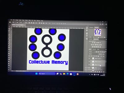 The image shows a computer screen displaying Adobe Photoshop software. On the screen, there is a design with blue circles and white rings on a white background. The text "Collective Memory" is visible in blue lettering below the design. The Photoshop interface shows various panels, including Layers, Adjustments, and Navigator. The computer's taskbar is visible at the bottom of the screen, displaying the time as 19:48 and the date 17/06/2026. The scene is a dimly lit room, with the glow of the computer screen being the primary light source. There is no indication of the city of Mararaba, Nigeria, or any people in the image.