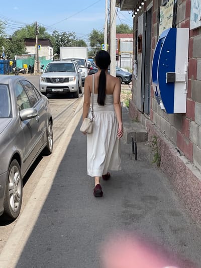 A woman with long black hair in a ponytail, wearing a strapless cream-colored dress and burgundy loafers, walks away from the camera on a sidewalk next to a brick building. She carries a small cream-colored purse. To her left, a silver Mercedes-Benz is parked. Further down the street, a silver Nissan SUV and a white truck are in traffic. The building to her right has a blue and white public telephone on its exterior wall. The scene appears to be in a commercial or residential area in Almaty, Kazakhstan, as indicated by the license plate on the Nissan SUV, which reads "792 BHF 05" and resembles Kazakh plates. The sky is clear and blue, suggesting a sunny day.