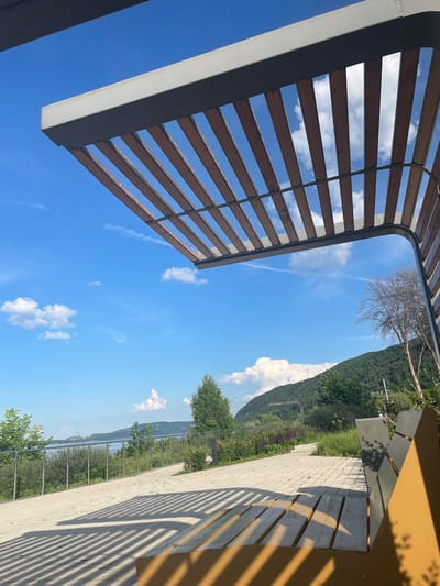 This is a wide-angle, outdoor, eye-level shot taken on a clear day in Shiryaevo, Russia. The scene features a modern, open-air shelter with a slatted wooden roof and a metal frame, casting striped shadows on the paved ground below. A yellow and grey park bench is visible in the foreground, partially obscured by shadows and the shelter's structure. Beyond the paved area, a metal railing lines a walkway, separating it from lush green vegetation and trees. In the background, a body of water, likely the Volga River, stretches towards rolling hills covered in dense forest. The sky is a vibrant blue with scattered white clouds. The image captures a serene, park-like environment overlooking a natural landscape.