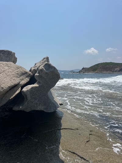 The image depicts a rocky coastline on a clear, sunny day. In the foreground, large, weathered grey boulders are positioned on a sandy beach. The rocks have intricate shapes and shadows cast by the sun. The sea is visible in the mid-ground, with waves breaking and creating white foam as they approach the shore. In the distance, a lush green hill rises from the sea, with a small structure, possibly a lighthouse or monument, visible at its peak. The sky is a bright, clear blue with a few wispy white clouds. The scene suggests a natural, somewhat wild coastal environment. This location is identified as being in Castiadas, Italy.
