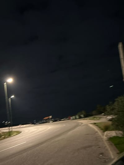 The image is a night photograph depicting an outdoor street scene, likely in Anatoliki Attiki, Greece. The perspective looks along a gently curving asphalt road, which occupies the right half of the frame. A white line marking is visible on the road surface, illuminated by artificial light.

The sky is dark, characteristic of nighttime, with faint, darker cloud-like patches visible in the upper left. High in the sky, a thin, bright, linear light streak is discernible, possibly a satellite or a distant aircraft light trail.

Two prominent streetlights illuminate the scene from the lower-central part of the image. The closer, brighter streetlight casts significant light on the foreground. Along the right side of the road, vegetation including trees, bushes, and grassy areas are visible. In the distance, along the road's edge, some indistinct structures or buildings can be faintly seen.

No people are present in the image, and no specific activity or event is taking place. The weather appears clear at night. There is no visible text.