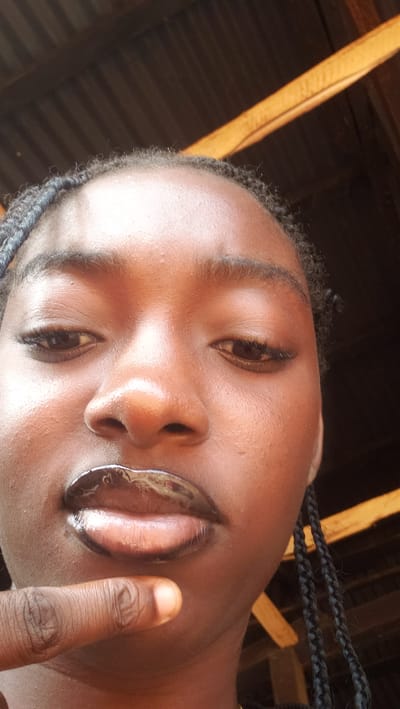 The media file is a close-up, low-angle selfie of a person's face, tilted to the right. The person has dark skin and is wearing braided hair that is visible on the sides of their head. Their eyes are open and looking slightly upwards and to the left. Their lips are parted and appear glossy, possibly due to lip gloss or balm. A finger is visible on the right side of the image, touching the person's cheek or chin area.
The background suggests an indoor setting, possibly a room with a wooden ceiling and diagonal wooden beams. The lighting appears to be natural and somewhat dim. There are no visible signs of text or writing. The overall impression is a casual, personal snapshot. The location context provided is "Television, Nigeria," which is unusual and could refer to a television program being filmed, a set designed to resemble a Nigerian environment, or a reference to media within Nigeria. Without further context, it's difficult to ascertain the exact nature of this location. The image does not convey a specific time of day or weather conditions, and the emotion conveyed is neutral to perhaps playful due to the finger gesture.