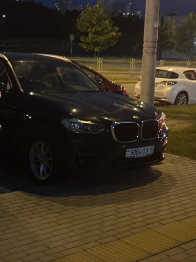 The image shows a black BMW parked on a paved area at night. The car's license plate reads "9064 OX-5". In the background, there are trees, streetlights, and buildings, indicating an urban or suburban environment. A white car and a red car are also partially visible. The paved area appears to be a sidewalk or parking lot, with a distinct tactile paving strip at the bottom right. The overall lighting suggests it is nighttime. The location is identified as Baravliany Village Council, Belarus, likely a street or public space within this area.