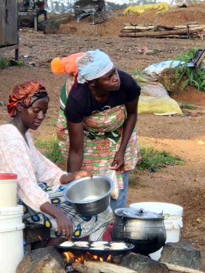 The image depicts an outdoor scene in Kabong, Nigeria, where two women are preparing food over an open fire. In the foreground, a woman wearing a patterned headscarf and a light-colored, floral-print top is seated. She is actively engaged in cooking, holding a metal bowl in her right hand and tending to small, flat, pale-colored items cooking on a griddle over flames. Beside her, to her left, are stacked white containers with red lids.
Behind her, another woman, also wearing a headscarf and a patterned apron over a dark top, stands and observes. Her hands are partially visible. The cooking area features a griddle placed on rocks over burning wood. To the right of the standing woman, large metal pots are visible, one with a lid.
The environment appears to be a dusty, unpaved area with sparse vegetation. In the background, there are piles of dirt, what appear to be logs or building materials, and large sacks, suggesting a possibly industrial or market setting. The overall impression is one of daily life and food preparation in an outdoor, possibly informal, setting.