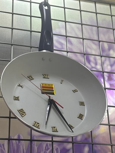A frying pan clock hangs on a wall with a grid-like design overlaying a purple floral pattern. The clock face is white with gold Roman numerals and a black handle. The time displayed is approximately 1:12. The clock has a logo near the center that includes the word "VARI". There is no evidence to confirm the city of Klin, Russia.