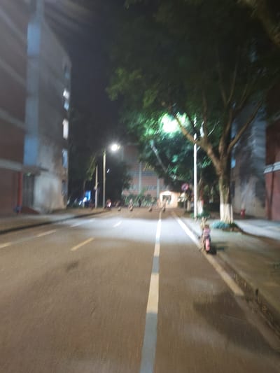 The image depicts a blurred nighttime street scene. A multi-story building, possibly an apartment complex or dormitory, is visible on the left side of the street, featuring multiple windows. Across the street, another building is partially visible on the right, with trees lining the sidewalk. A street lamp illuminates the area. A pink-colored scooter is parked on the right sidewalk. Several figures, appearing as blurred shapes, are present in the distance, some near the scooter and others further down the street, suggesting pedestrian or vehicular activity. The road is paved with asphalt and marked with white and yellow lines. The overall impression is of a street in a residential or campus area at night, with the blur indicating motion or low light conditions.