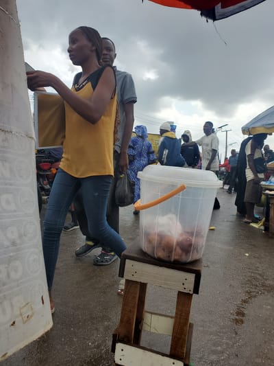 The image depicts a bustling outdoor market scene in Gwong, Nigeria, under a cloudy sky. The foreground is dominated by a clear plastic bucket filled with round, brown food items, likely fritters or dumplings, resting on a wooden stool. To the left, a young woman with braided hair, wearing a yellow and black sleeveless top and blue jeans, is seen from the side, appearing to interact with something off-camera. Behind her, a man in a gray t-shirt looks towards her.
In the mid-ground, several other individuals are visible. A person in a blue and white patterned garment is walking away, holding a black plastic bag. Further back, a group of men are gathered, one wearing a white t-shirt, another in a hooded jacket. A red motorcycle is parked to the left of the woman.
The environment suggests a street market setting, with a wet, asphalt surface indicating recent rain. Various stalls and awnings are present in the background, along with overhead power lines. The overall atmosphere is one of commerce and daily life in a Nigerian urban setting.