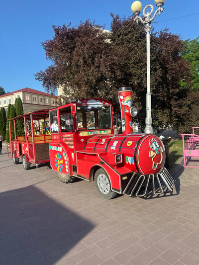 This media file depicts a small, bright red train designed for children's entertainment, parked on a paved sidewalk in Ternopil, Ukraine. The train is elaborately decorated with cartoon characters and colorful decals, including clouds, rainbows, and smiling faces. It appears to be a tourist attraction or a recreational vehicle for a park.
The train's front section resembles a locomotive, complete with a smokestack featuring a rainbow design and a headlight. A driver's cabin with large windows is visible, suggesting it's a functional vehicle. Behind the cabin are open-air passenger cars with railings, ready to accommodate children. The train is adorned with Cyrillic text, though it is partially obscured or difficult to read clearly in the image. Some visible words include "ВЕСЕЛЫЙ" (Veselyy - Merry) and possibly "ПОТЯГ" (Potyah - Train) on the side.
The setting is outdoors, with a clear blue sky indicating a sunny day. Lush green trees and a building with a red roof and numerous windows are visible in the background, providing context for an urban or park-like environment. A street lamp with ornate details stands beside the train. The overall impression is cheerful and inviting, typical of a family-friendly attraction. The time of day appears to be during daylight hours, with the sun casting shadows on the pavement. No people are visible in the immediate vicinity of the train.