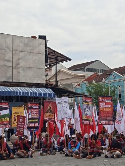 A group of people are seated on the ground in front of a large building with a white, tiled facade. They are holding up large signs with Indonesian text and graphics, which are mostly red and white. There is a large billboard behind the protesters. The image is taken from a slightly elevated angle. The location is on a street in Yogyakarta, Indonesia, based on the coordinates. The people are wearing casual clothes and some are wearing orange vests. The atmosphere is tense and the signs suggest a protest. The image shows a demonstration in progress.