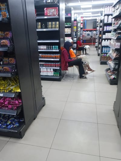 The image displays an indoor commercial retail establishment, located in Kawaji, Nigeria. The facility features aisles of uniformly black shelving units stocked with various consumer products, set on a floor of light-colored square tiles with dark grout lines. Overhead fluorescent lighting illuminates the entire space.
In the mid-ground, three individuals are seated on stools. On the left, a person, likely female, wears a dark blue hijab, a maroon long-sleeved top, and black trousers, seated on a metal stool and looking downwards. Next to her, a second person, also likely female, wears an orange long-sleeved top and a patterned lower garment, seated on a similar metal stool and looking downwards; she wears a white slip-on sandal on one foot, with the other foot appearing bare or in very light footwear. Further back and to the right, a third person, likely female, with short hair, wears a red short-sleeved top and dark trousers, seated on a low stool while appearing to engage with an item on a small white table positioned in front of her.
The shelving unit in the left foreground prominently displays multiple packages of "FOX'S Chocolately BIG CHUNKY COOKIES," along with shelves containing numerous packages of yellow, pink, and blue-wrapped items, consistent with snacks or confectionery. The shelving units in the right mid-ground are stocked with personal care products, including perfume gift sets, one labeled "SCARLET KISS," various boxed fragrances, and multiple rows of aerosol cans such as deodorants, with some products from the brand "COSMO" visible. The aisles extend into the background, revealing additional stocked shelving and a structural racking system in blue and red.