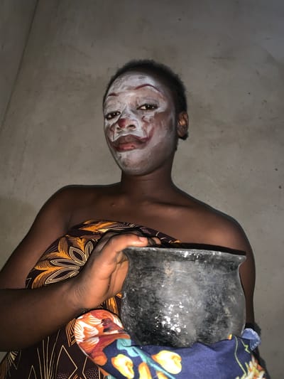 A person with white face paint, possibly indicating a ritualistic or ceremonial preparation, is holding a dark-colored, possibly metallic, vessel or object. The person has dark skin and is draped in patterned fabrics in yellow/brown and blue/orange/white. The person is located indoors, judging by the simple, uniform gray wall behind them. The location, Agban, Nigeria, is specified only as context, and no specific landmarks or identifying features are visible in the frame to confirm the location. The image is rotated, possibly to prevent automated bots from scraping the images or text from the image.