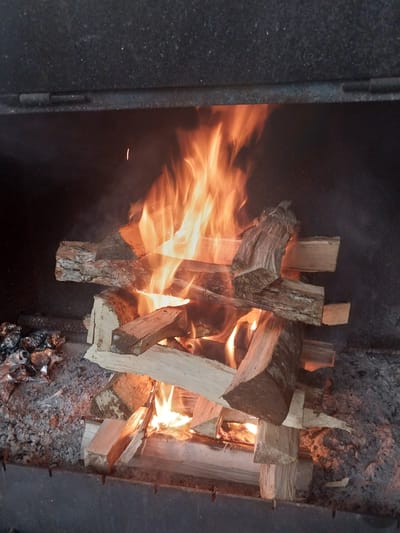 This media file shows a close-up, overhead view of a fire burning in what appears to be a fireplace or fire pit. The main subjects are logs of firewood, some of which are actively burning with bright orange flames. The fire is the central focus, with flames licking and swirling around the wood. There is also smoke visible, rising from the fire and obscuring some of the background.

The scene is set within a dark enclosure, with textured, dark gray walls on the left and right sides of the image. These walls suggest the confines of a firebox or an outdoor structure. The ground beneath the fire is covered in ash and embers, with some larger pieces of charred wood or debris visible towards the bottom right. The overall impression is one of intense heat and combustion.

The activity taking place is a fire burning. The logs are arranged in a way that suggests they are meant to combust, possibly for warmth or cooking. The flames are dynamic and appear to be consuming the wood.

Notable details include the texture of the firewood, which varies from rough bark to smoother, cut surfaces. The light from the fire illuminates the wood and creates strong contrasts. There is no visible text in the image. Given the location context of Kapan, Armenia, this could be a traditional hearth or an outdoor fire used for heating or preparing food. The time of day and weather are not discernible from the image. The emotion conveyed is primarily one of raw energy and heat.