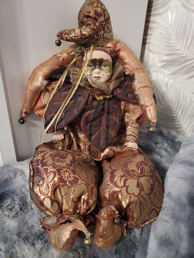 The image shows a porcelain jester doll with a mask. The doll is dressed in a detailed costume of gold and maroon brocade fabric, with a ruffled collar and bell-tipped points on its hat. The doll's face has painted features, including blue eyes and a neutral expression. It is lying on a white surface, with a grey furry material in the background. A patterned wallpaper with leaf motifs is also visible behind the doll. The overall impression is that of a decorative collectible item. There is no text visible in the image. The context is Tiraspol, Moldova.