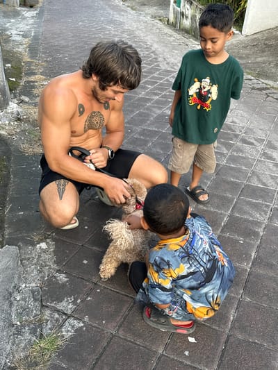 Captured from an elevated, oblique angle in what appears to be Dalung, Indonesia, the scene features an adult male, two young boys, and a poodle. The environment consists of a paved area constructed from interlocking gray tiles, interspersed with patches of grass and moss along the edges, indicative of an outdoor setting in a tropical or subtropical climate.
The adult male is shirtless, wearing black athletic shorts, a watch on his left wrist, and sandals on his feet. He has tattoos on his chest and right thigh. He is crouching down and holds a leash.
Two young boys stand near him. One boy wears a green t-shirt with a graphic design, khaki shorts, and sandals. The other is wearing a brightly colored shirt with orange and yellow floral patters, athletic-style footwear.
The poodle is centrally located, held or petted by the adult.
The photograph's focus and framing suggest candid interaction. The perspective is from an elevated point, giving a view of the subjects.