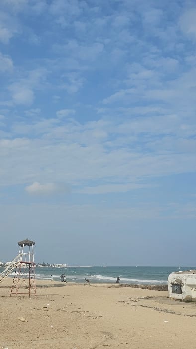 This image depicts a beach scene in Sousse, Tunisia, under a partially cloudy blue sky. In the foreground, a sandy beach extends towards the Mediterranean Sea. To the left, a weathered metal lifeguard stand with a white ladder and a dark, possibly thatched, roof is present. To the right, a dilapidated, light-colored concrete structure with a rectangular opening is visible, partially buried in the sand. A low, stone or concrete wall or barrier runs horizontally across the mid-ground.

Several individuals are present on the beach near the waterline. One figure on the far left of the group appears to be in or near the water, possibly wading. Other figures are standing or walking along the shoreline. The sea exhibits moderate waves, appearing blue-green, and distant vessels or structures are discernible on the horizon. The coastline in the distance shows faint outlines of urban development, including buildings. Debris and footprints are scattered across the sand.