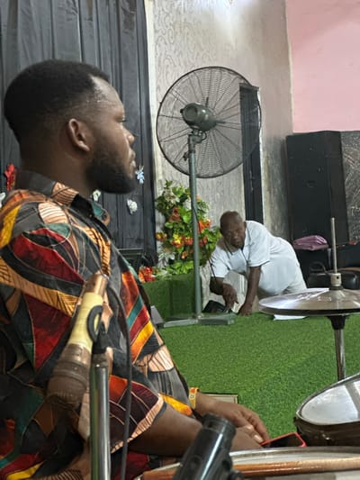 The scene takes place in what appears to be a room in Aba, Nigeria, as indicated by contextual clues. A man in a colorful patterned shirt and beard is positioned in the foreground, with his head tilted upwards. A microphone is visible near him, suggesting a possible performance or recording activity. Behind him, another man wearing a white shirt is kneeling, possibly interacting with some equipment or items on the floor, which is partly covered with a patch of artificial grass. A black fan and a flower arrangement are also present in the background. The room has a tiled floor and the walls are painted in light colors.