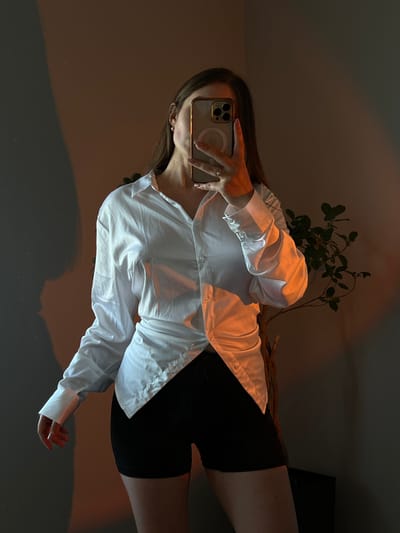 The image is a selfie taken by a person in Ternopil, Ukraine. The person is wearing a white button-down shirt and black shorts. They are holding a smartphone in their right hand, using it to take the picture in a mirror. A plant with green leaves is visible behind the person. The lighting in the room is dim, with warm-toned light casting shadows. The overall mood appears to be relaxed and casual. The time of day is not explicitly indicated, but the lighting suggests it could be evening or indoors with artificial lighting. The background is mostly dark, with some hints of furniture or other objects not clearly discernible. There is no visible text in the image.