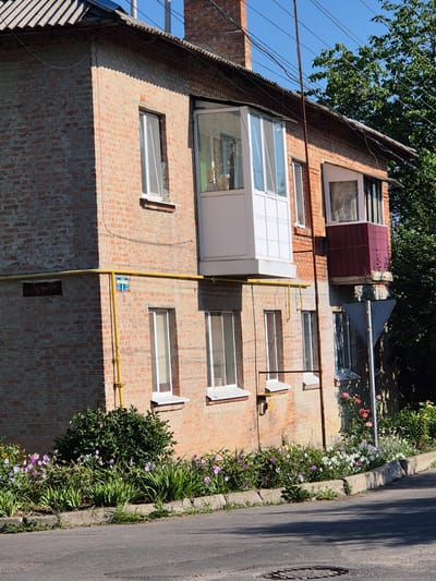 The image depicts a brick building located in Sozonivka, Ukraine. The building has multiple windows and two balconies, one of which is enclosed with glass. A yellow pipe runs along the exterior of the building, with a blue and white sign attached to it. In front of the building, there is a flowerbed with various types of flowers, including pink, purple, and white blooms, as well as green foliage. A concrete curb separates the flowerbed from the asphalt road. Trees are visible in the background. The sky is clear and blue. The scene is illuminated by sunlight, casting shadows on the ground.