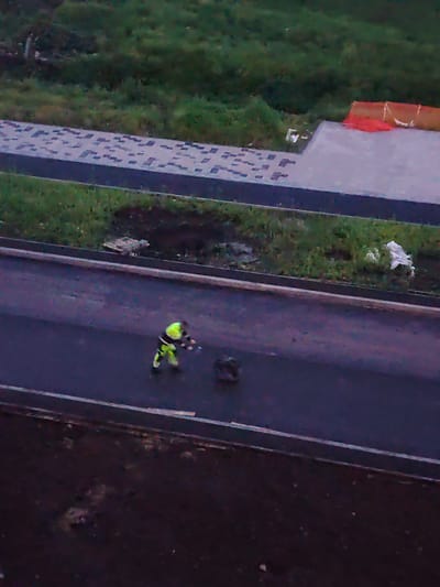 An aerial view shows a person in a high-visibility yellow and green outfit working on an asphalt road. The individual is holding what appears to be a tool or equipment in their hands. To the right of the worker, a dark, circular object is visible on the road. Adjacent to the road is a grassy area with some debris, and further beyond is a paved walkway with a pattern of darker squares. In the background, a large area of green vegetation is visible. The scene appears to be an outdoor construction or maintenance site in Yekaterinburg, Russia.