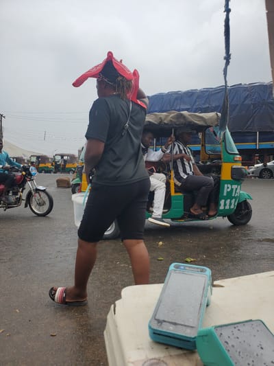 The image depicts a street scene in Gwong, Nigeria. The focal point is a person wearing a black t-shirt, black shorts, a red bandana on their head, and sandals. Their right leg is extended, and they are looking downwards. Behind this person, a yellow and green auto-rickshaw, also known as a "keke," is visible. Two other individuals are seated within the keke. One is wearing a black and white striped shirt and black pants, holding a metal rod. The other is wearing a white shirt and pants. The keke has "PTA 1111" written on its front.
Further back, a line of similar auto-rickshaws is parked. In the foreground, to the right of the main subject, are two turquoise electronic devices, possibly mobile phones or payment terminals, with screens displaying a grid-like pattern and water droplets. The ground is wet, suggesting recent rain or a damp environment. The sky is overcast, indicating a cloudy day. The scene appears to be outdoors, possibly a market or a transportation hub, given the presence of multiple vehicles. The overall atmosphere seems casual, with individuals engaged in daily activities.
