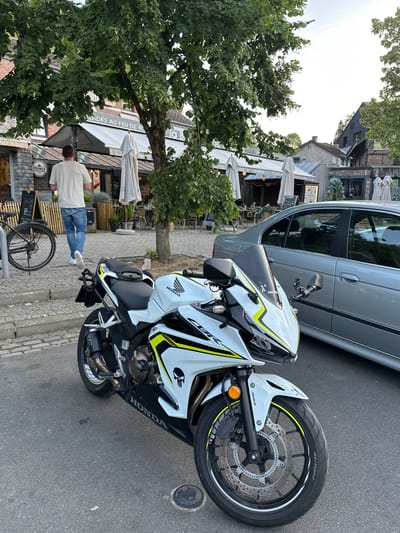 In Durbuy, Belgium, a white Honda CBR motorcycle with yellow accents and a skull decal is parked on a cobblestone street. The motorcycle is in the foreground, angled towards the right. A silver sedan is parked to the right of the motorcycle. In the background, a man wearing a light-colored t-shirt and blue jeans walks away from the camera. He is near a bicycle parked on the sidewalk. A building with an awning and outdoor seating is visible behind the man. Trees with green leaves are overhead, and the sky is bright, suggesting daytime.