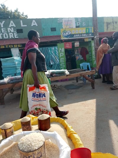 The image shows a street scene in Malava, Kenya, located at latitude 0.489 and longitude 34.837. A woman is walking down the street, carrying a large bag of rice. She is walking past a store with a sign that reads "JUMILA & SMILE SM."  The woman is wearing a green skirt and a red and black shawl. She is holding a bag of "Kinrie" rice. The street is paved and there is a small plastic chair on the sidewalk. There are several bags of white rice in the foreground, which are on a yellow container.  The image was taken in a sunny, daylight environment.  The store is brightly colored, painted in shades of green and yellow.  The woman is walking to the right of the frame.  The building behind her is decorated with a large, yellow sign that reads "Fanta."  The store appears to be in business.  The street is not busy, and only the woman is in the foreground.  There are two people standing on the other side of the street in the distance.  They are a man and a woman.  The woman is wearing a long purple dress and a brown shawl.  The man is wearing blue jeans and a black shirt.  They appear to be talking.  They are out of focus, and the woman in the foreground is the subject of the image.  The woman carrying the bag of rice is walking toward the store.  There is a tree in the background.  The weather appears to be good, with a clear sky.  There are no other vehicles or pedestrians visible.  The image is taken from a low angle, looking up at the woman walking.