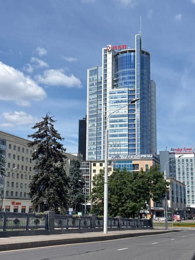 The image shows a daytime view of a city street in Minsk, Belarus. A tall, modern skyscraper with a curved glass facade dominates the skyline. Red lettering on the building reads "BSB" and "ТЕХНОБАНК". To the right, another building with red lettering says "АЛЬФА-БАНК". In the foreground, there is a street with a sidewalk and a decorative metal fence. Several trees, including a large pine tree, are visible. The sky is blue with scattered white clouds.