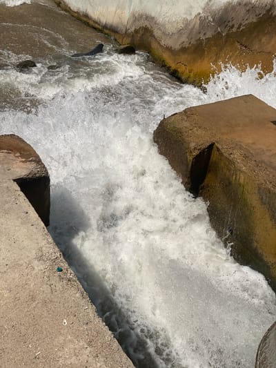 This media file captures a close-up view of water cascading over a concrete structure, likely a spillway or dam. The water is turbulent and foamy, indicating a strong flow. The surrounding environment is industrial, with concrete channels and rough-hewn concrete or stone elements visible. Some of these elements appear aged, with some moss or algae growth visible on the wetter surfaces. The lighting suggests it is daytime, with bright sunlight casting shadows.

The scene is dominated by the dynamic movement of water, which appears to be rushing downwards. The concrete structures are angular and functional, designed to control or channel the water. There are no people visible in the frame. The presence of what appears to be cigarette butts and a small green object on the concrete surface in the lower right of the image suggest human activity in the vicinity, though not directly in the water.

The overall impression is one of raw, natural power being managed by man-made infrastructure. The water's frothy surface and the shadows suggest a sunny day. The setting could be an urban waterway, a hydroelectric facility, or an irrigation system. Without further context, it's difficult to pinpoint the exact location beyond the general industrial and water-control theme. The image does not contain any visible text.