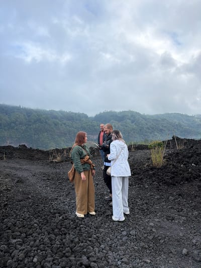 The image shows a group of four people standing on a landscape of dark volcanic rocks under a cloudy sky. In the foreground, a woman with red hair, wearing a green jacket and brown pants, is looking to her left. Beside her, a man in a black jacket and light-colored pants is looking at a phone. To their right, a woman in a white jacket and light-colored pants is facing away from the camera. In the background, a man in a red shirt stands behind the other three. The terrain is covered with small, dark, porous volcanic rocks. Beyond the rocks, a lush, green, forested mountainside rises up, shrouded in mist. The scene appears to be the rim of a volcanic crater or a recent lava field, consistent with the geological features found in Kintamani, Indonesia, a region known for its active volcanoes and crater lakes. The overall atmosphere is overcast and somewhat damp, suggesting a cool, possibly humid, climate.
