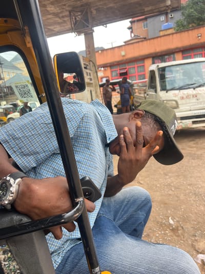 A medium shot captures a Black man sitting in the driver's seat of a yellow auto-rickshaw in Nkpor, Nigeria. He is wearing a blue and white checkered shirt, a green baseball cap, and blue jeans. His right hand is raised, covering his face as if shielding his eyes from the sun or posing for the photo. A black smartphone is held in his left hand, resting on his lap. A silver watch with a black band is visible on his left wrist.
The background depicts a bustling street scene with buildings and vehicles. To the left, a white truck with red accents and text is partially visible. A building with orange and red facade and multiple windows is also present. The ground is sandy and appears dry, suggesting a sunny day. A side-view mirror of the auto-rickshaw is in the foreground, reflecting the surroundings, including a glimpse of the driver's face and the scene behind. A QR code sticker is affixed to the windshield. The overall impression is that of a typical day in a busy Nigerian urban environment. The man's gesture of covering his face could suggest a desire for privacy or a playful interaction with the photographer.