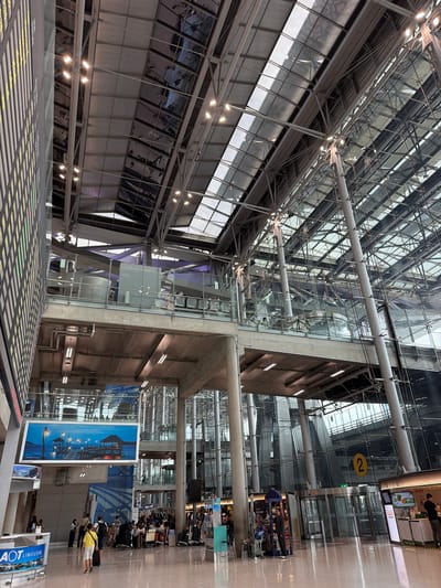 The image shows the interior of a large, modern airport terminal, likely Suvarnabhumi Airport, located in Nong Prue Subdistrict, Thailand. The scene is characterized by its expansive, high-ceilinged architecture with a prominent use of glass and metal structures. Sunlight streams in through a large, multi-paneled glass roof and walls, reflecting off the polished floor.
On the left side of the image, a large electronic display board with rows of yellow and green text is visible, likely showing flight information. Below this, an "AOT Limousine" sign is displayed on a blue background with images of cars.
In the mid-ground, several people are seen walking and standing with luggage. There are seating areas and various service counters or shops. A prominent sign reads "LUGGAGE WRAPPING." A large framed picture depicting a tranquil scene of piers and huts at dusk is hung on a wall.
Further into the terminal, a large crowd of people with trolleys full of luggage suggests an active travel environment, likely near check-in counters or baggage claim. A yellow circle with the number "2" is prominently displayed on a pillar, possibly indicating a gate or section.
The architectural style emphasizes open spaces, transparency with extensive glass facades, and structural beams and columns supporting the roof and upper levels. The overall impression is one of a busy, functional, and architecturally impressive transportation hub.