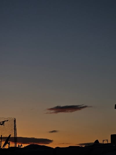 The image depicts a transitional sky, likely at dusk or dawn, over the city of Fez, Morocco. The upper sky is a dark blue-grey, gradually transitioning to orange and yellow hues at the horizon line. Several elongated, dark clouds are present, with one prominent cloud in the upper-middle exhibiting reddish-orange coloration on its underside. Silhouetted against the vibrant horizon on the left is a tall, dark structure resembling a chain-link fence, with a satellite dish visible below it. The silhouette of a city skyline stretches across the lower portion of the frame, showing uneven rooftops and various architectural structures, including what appears to be a small, open framework on a rooftop to the right. No individuals, specific actions, or direct interactions are discernible within the scene.
